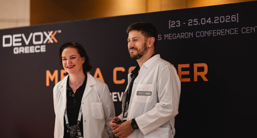 Marit van Dijk and Piotr Przybył in front of a Devoxx Greece banner; both are wearing a white labcoat.