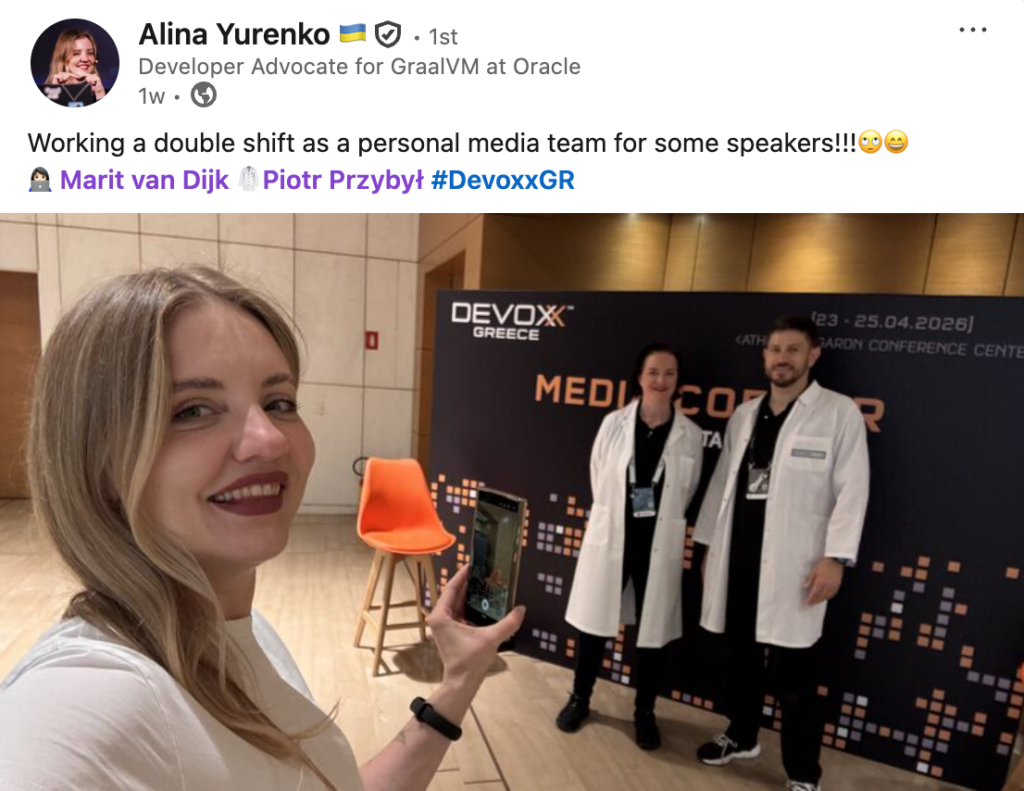 Social media post by Alina Yurenko with the text: "Working a double shift as a personal media team for some speakers!!!🙄😄" tagging Marit van Dijk, Piotr Przybył and Devoxx Greece. The post contains a selfie of Alina Yurenko. Marit van Dijk and Piotr Przybył are standing in the background in front of a Devoxx Greece banner; both are wearing a white labcoat.