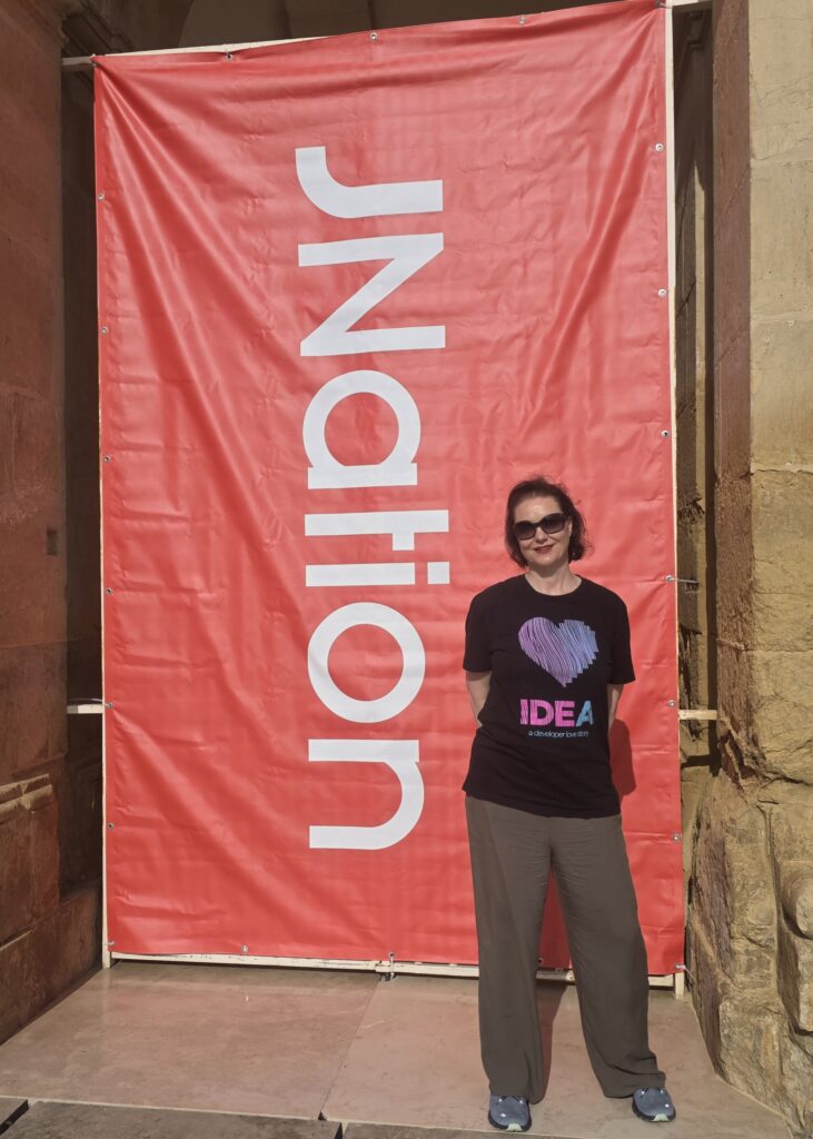 Marit van Dijk wearing a black IntelliJ IDEA t-shirt and sunglasses, standing in front of a huge red JNation banner.
