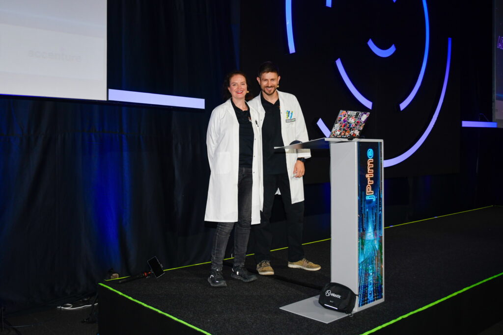 Marit van Dijk and Piotr Przybył posing for a picture on stage at jPrime. Both are wearing a labcoat.